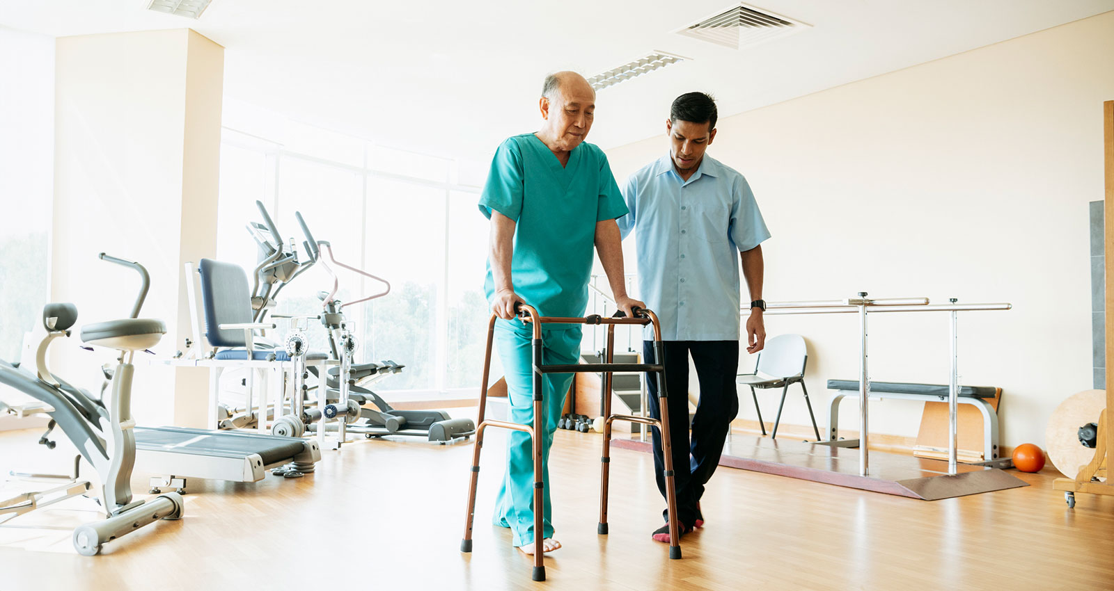 Male physiotherapist supporting Malaysian patient in his 70s, learning to walk with frame